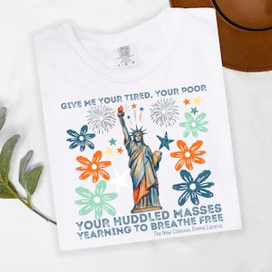 Puede incluir: Camiseta blanca con un dise&ntilde;o de la Estatua de la Libertad con fuegos artificiales y flores. La camiseta tiene el texto "GIVE ME YOUR TIRED, YOUR POOR" y "YOUR HUDDLED MASSES YEARNING TO BREATHE FREE".