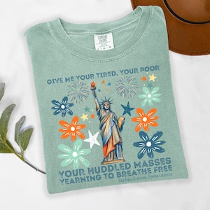 Puede incluir: Una camiseta verde salvia con un dise&ntilde;o de la Estatua de la Libertad con una antorcha, rodeada de flores y fuegos artificiales. El texto dice "GIVE ME YOUR TIRED, YOUR POOR, YOUR HUDDLED MASSES YEARNING TO BREATHE FREE" del poema de Emma Lazarus.