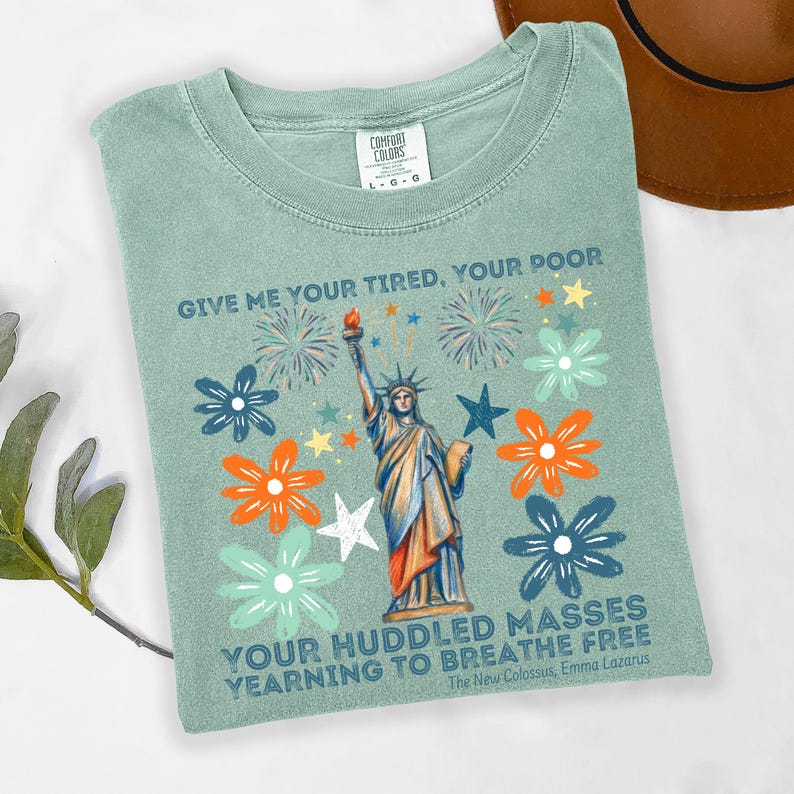 Puede incluir: Una camiseta verde salvia con un dise&ntilde;o de la Estatua de la Libertad con una antorcha, rodeada de flores y fuegos artificiales. El texto dice "GIVE ME YOUR TIRED, YOUR POOR, YOUR HUDDLED MASSES YEARNING TO BREATHE FREE" del poema de Emma Lazarus.