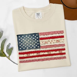 May include: Cream-colored t-shirt featuring a decorative American flag design. The flag's stars are on a denim-like patch, and the stripes are made of red gingham, floral, and lace fabrics. The shirt has a Comfort Colors tag.