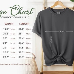 Puede incluir: Una camiseta gris oscuro exhibida en una percha de madera, con medidas de ancho y largo. Una tabla de tallas muestra las dimensiones en pulgadas y cent&iacute;metros para las tallas S a 4XL. El texto "Size Chart" y "Comfort Colors 1717" tambi&eacute;n son visibles.