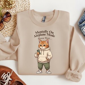 May include: Beige sweatshirt with a graphic of a cat wearing a hoodie and holding a phone. The text on the sweatshirt reads "Mentally On Airplane Mode, Please Wait..."