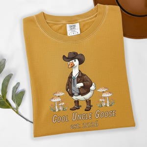 May include: Mustard yellow Comfort Colors t-shirt featuring a cartoon goose dressed as a cowboy. The goose wears a brown hat, jacket, and boots. Text reads "Cool Uncle Goose est. 2026".