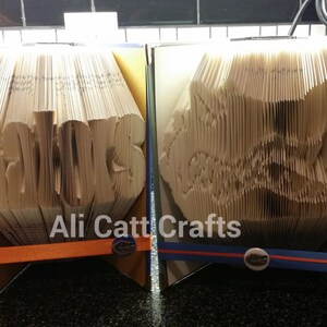 May include: Two book folding crafts, one with the word "Gators" and the other with a Gator head design. Both are made from old books and have a blue and orange ribbon.