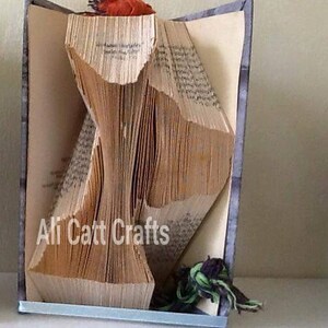 Pode incluir: Um livro dobrado na forma de um pássaro, com um detalhe em tecido vermelho e laranja no topo. O livro está em pé sobre uma mesa com um detalhe em tecido azul e verde na parte inferior. O texto "Ali Catt Crafts" é visível no livro.