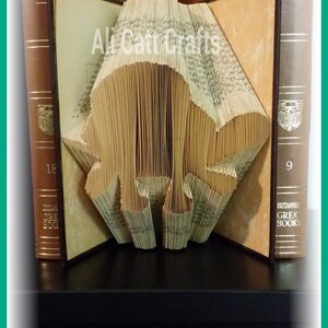 May include: A book folded into the shape of a bear, standing on a black surface between two brown books. The book on the left has the text "Augustus" and the book on the right has the text "Aristotle II".