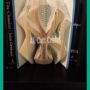 May include: A book art sculpture of a tree, folded from the pages of a book. The sculpture is displayed on a shelf with two other books, one titled "The Chamber" by John Grisham and the other titled "Eclipse" by Stephenie Meyer.