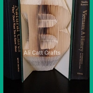 May include: A book folded into the shape of the letter "B" with the title "Natural Cures" on the left side and "Vietnam: A History" on the right side. The book is sitting on a black surface.