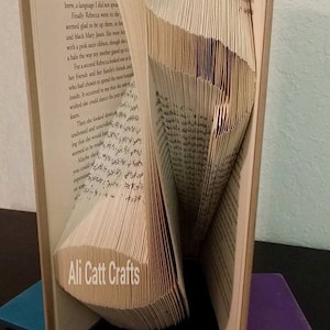 May include: A book folded into a three-dimensional shape, creating a sculpture of a bird. The book is open and the pages are folded in a complex pattern. The book is sitting on a table with a blue and purple bookend on either side.