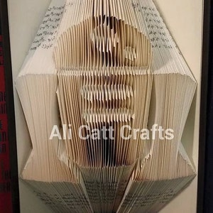 May include: A book sculpture with pages folded to create a detailed silhouette of a person. The artwork is displayed open, revealing the intricate folds and the text of the book. The artist's name, "Ali Catt Crafts," is visible.