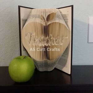 May include: A book folded into the shape of an apple with the word "Teacher" written inside. The apple is sitting on a black surface next to a green apple. The text "Ali Catt Crafts" is printed below the apple.
