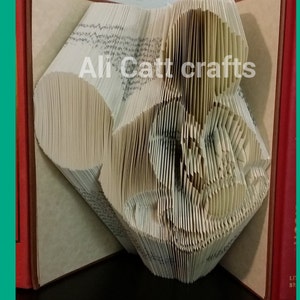 May include: A book folded into the shape of a cat's head. The book is open and the pages are folded to create the shape of the cat's head. The book is sitting on a black surface. The text "Ali Catt crafts" is visible on the book.
