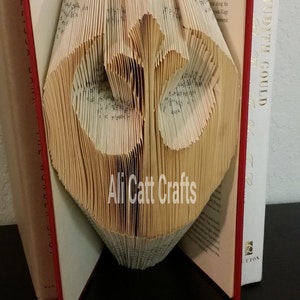 May include: A book art sculpture of a heart shape made from the pages of a book. The book is open and the pages are folded into a three-dimensional heart shape. The book is red and has the text "Ali Catt Crafts" on the pages.