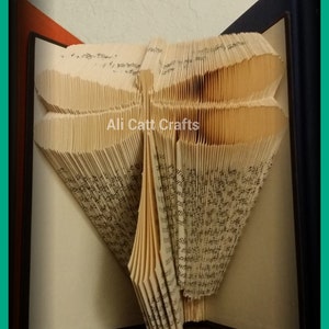 May include: A book art sculpture of a dragonfly, folded from the pages of a book. The sculpture is displayed on a shelf with a dark brown surface. The book art is titled "Ali Catt Crafts".