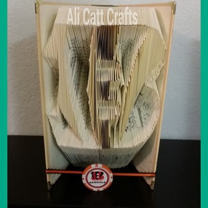 Könnte beinhalten: Ein Buch, das in die Form der Zahl "12" gefaltet ist, mit einem Cincinnati Bengals-Logo auf der Vorderseite. Das Buch liegt auf einer dunklen Oberfläche.