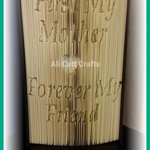 Könnte beinhalten: Ein beigefarbenes Buch mit den Worten "First My Mom" und "Forever My Friend", die in die Titelseite geschnitzt sind. Das Buch ist geschlossen und steht aufrecht auf einer schwarzen Oberfläche. Ein Herz ist in das Buch geschnitzt, darunter die Worte "Ali Catt Crafts".