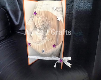 Può includere: Un libro piegato a forma di lettera C, decorato con stelle viola e argentate. Il libro è in piedi su una superficie nera e ha un nastro argentato legato intorno.