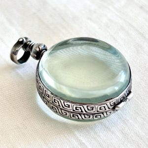 Può includere: Un ciondolo rotondo argentato con un globo di vetro trasparente verde chiaro. Il ciondolo ha un bordo decorativo con un motivo geometrico ripetuto. L'anello è un semplice anello attaccato a una parte superiore decorativa.