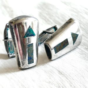 May include: A pair of silver cufflinks with geometric designs. Each cufflink features a rectangular and triangular inlay of teal and blue-green stone. The cufflinks are set against a white background.