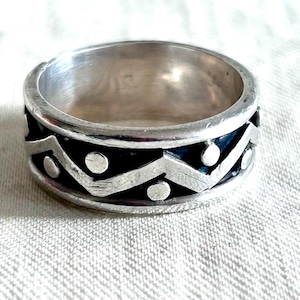 May include: A silver ring with a geometric design. The ring features a band with alternating zigzags and circular accents. The design is set against a dark background, creating a contrast with the silver metal.