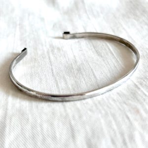 May include: A silver-colored cuff bracelet with a simple, open design. The bracelet is made of a thin, curved metal band. The ends of the bracelet are slightly flattened and bent. The bracelet is resting on a white, textured surface.