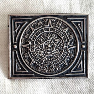 Può includere: Un ciondolo in metallo color argento con un designo in rilievo dettagliato del calendario azteco, noto anche come Pietra del Sole, circondato da una cornice quadrata.