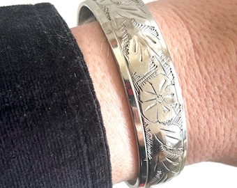 May include: Silver-toned bangle bracelet with an engraved floral design. The bracelet is wide and has a detailed pattern of flowers and leaves. The metal has a polished finish, reflecting light. The bracelet is worn on a wrist.