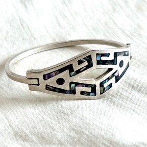 Könnte beinhalten: Ein silbernes Armreif-Armband mit einem geometrischen Design, das mit irisierendem Perlmutt eingelegt ist. Das Armband ist offen und hat einen Scharnierverschluss.
