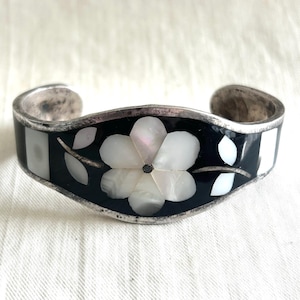 May include: A silver cuff bracelet featuring a central floral inlay made of iridescent white mother-of-pearl petals set against a smooth black background. The design includes small white leaf-shaped inlays and a metallic stem on each side.