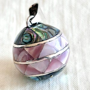 May include: A spherical pendant with a silver bail. The pendant is adorned with iridescent abalone shell and pink and lavender mother-of-pearl pieces, separated by silver bands. The overall design is geometric and eye-catching, suitable for jewelry.
