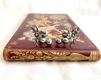 May include: A pair of silver earrings with a floral design and small round beads. The earrings are on a brown book with gold accents.