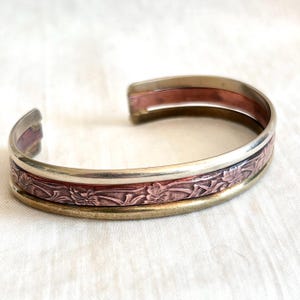 Op de afbeelding: Een manchetarmband van zilver, koper en messing. De armband heeft een bloemenpatroon in het midden, met een zilveren band erboven en een messing band eronder. De koperen band is aan de binnenkant van de armband zichtbaar.