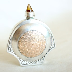 May include: A silver and gold Aztec calendar-themed perfume bottle with a clear amber stopper.
