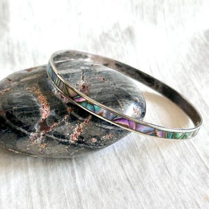 May include: A silver bangle bracelet with iridescent abalone shell inlay. The bracelet is resting on a dark gray stone. The abalone shell features shades of purple, green, and blue.