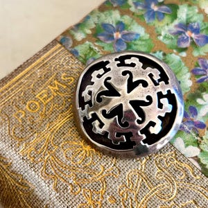 May include: A silver circular pendant with a black background and a stylized design of a four-pointed star.