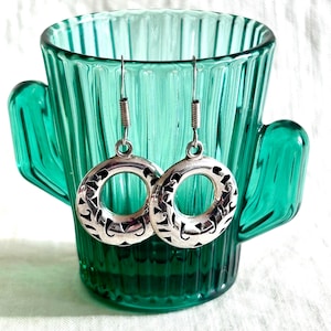 May include: A pair of silver-toned hoop earrings with intricate cut-out designs. The earrings are suspended from thin wires with coiled springs. The earrings are displayed in front of a green glass cactus-shaped container.