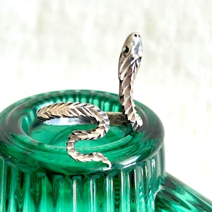 May include: A silver-toned metal ring shaped like a coiled snake. The ring features a textured, faceted surface that mimics scales along the body and head of the serpent. The design is open-ended, allowing the snake's head to rise upward.