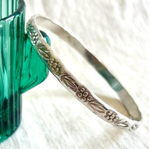 May include: A silver-toned metal bangle bracelet featuring a repeating embossed pattern of small circular clusters and elongated leaf-like shapes along the outer surface. The bracelet has a slightly irregular, hammered texture and a matte finish.