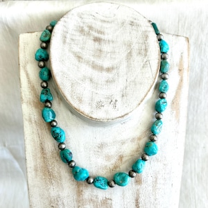 May include: A turquoise beaded necklace with silver-colored accent beads. The necklace is displayed on a white wooden jewelry stand. The turquoise beads are irregularly shaped and vary in size, creating a unique, handcrafted look.