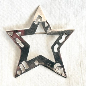 Könnte beinhalten: Eine silberne Brosche in Sternform mit einem Ausschnittmuster. Der Stern hat eine glatte, metallische Oberfläche und ist ein schlichtes, elegantes Schmuckstück.