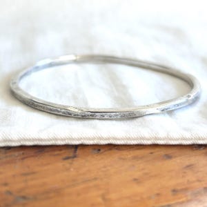 Op de afbeelding: Een zilverkleurige armband met een gehamerde textuur. De cirkelvormige armband lijkt van metaal te zijn. Het ligt op een witte doek, met een houten oppervlak op de achtergrond. De diameter van de armband is ongeveer 6,5 cm.