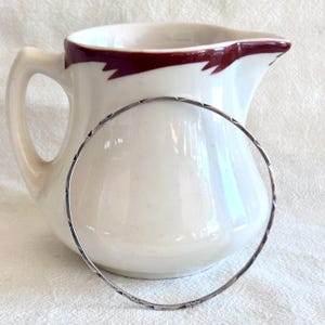 May include: A silver bangle bracelet with a textured design rests in front of a white ceramic pitcher with a maroon rim. The bracelet has a simple, elegant design. The pitcher is smooth and glossy.