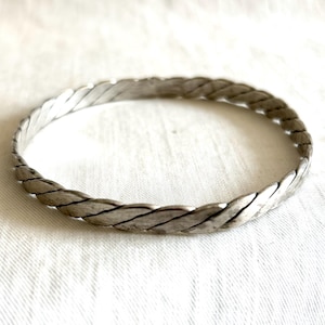May include: A silver-toned metal bangle bracelet featuring a continuous twisted rope design. The surface of the bracelet has a matte, slightly weathered finish with visible diagonal grooves that create a textured, braided appearance throughout the band.
