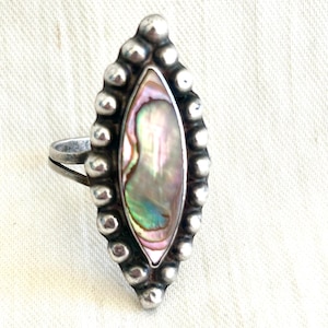 May include: A marquise-shaped ring featuring a central iridescent abalone shell inlay with pink, green, and blue tones. The stone is surrounded by a border of small, rounded silver beads, set on a simple, thin, polished silver band.