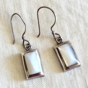 May include: A pair of drop earrings featuring rectangular silver settings that frame faceted white mother-of-pearl stones. Each earring is suspended from a curved silver ear wire, showcasing a polished metallic finish and a smooth, iridescent stone surface.