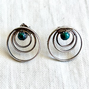 May include: A pair of silver-toned earrings with a circular design. Each earring features three concentric circles and a small, round, teal-colored stone at the center. The earrings are set against a white, textured background.