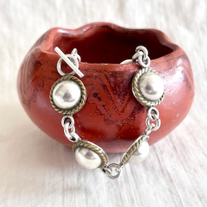 May include: A silver bracelet with large, round, polished silver beads. Each bead is framed with a gold-toned rope design. The bracelet is connected by silver links and has a toggle clasp. The bracelet is displayed in front of a red ceramic bowl.