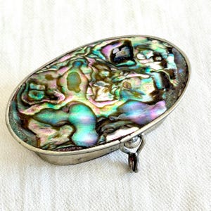 May include: Oval-shaped, iridescent abalone shell jewelry box with a silver-toned metal frame and hinge. The shell displays a vibrant mix of blues, greens, and purples. The box is closed, showcasing the polished surface of the shell.