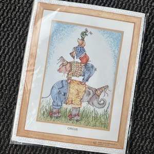 Op de afbeelding: Ingelijste kruissteekkunst met een circustafereel. Een olifant, een varken en een hond zijn op elkaar gestapeld en dragen feesthoeden en een spijkerbroek. Een kikker balanceert bovenop. Het woord "CIRCUS" staat onder de afbeelding.