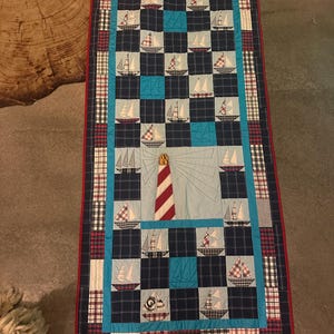 Pode incluir: Um caminho de mesa retangular com um design de patchwork de veleiros e um farol central. A colcha é composta por quadrados de tecido azul-marinho, azul-claro e xadrez, contornada por um debrum vermelho e uma moldura de tecido xadrez.